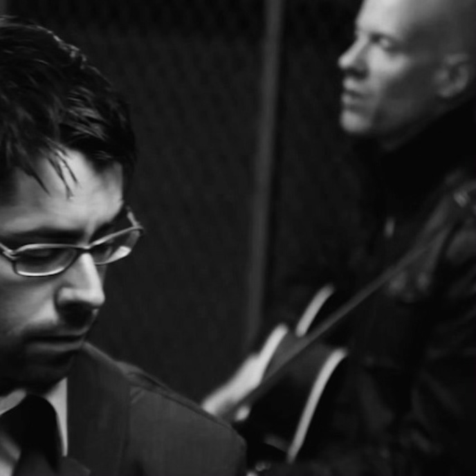 A black and white photo of the synth-pop duo Hot Coma. Multi-instrumentalist-producer Adam Boose is foregrounded and singer-songwriter-guitarist Robert Cherry is in the back of the photo, to the right. 